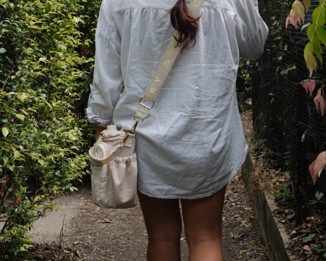 Person walking outdoors with a braided hairstyle, wearing a light-colored shirt and carrying a beige bag.